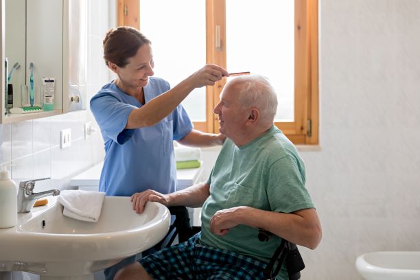 Older man in wheelchair in bathroom with care worker who is brushing his hair